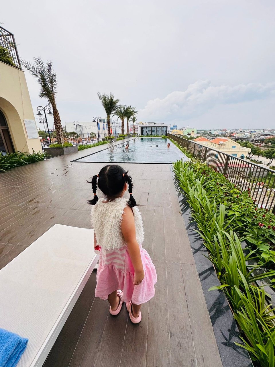 Hồ Bảo Tú mặc váy hồng đứng cạnh hồ bơi trên sân thượng, nhìn ra xa